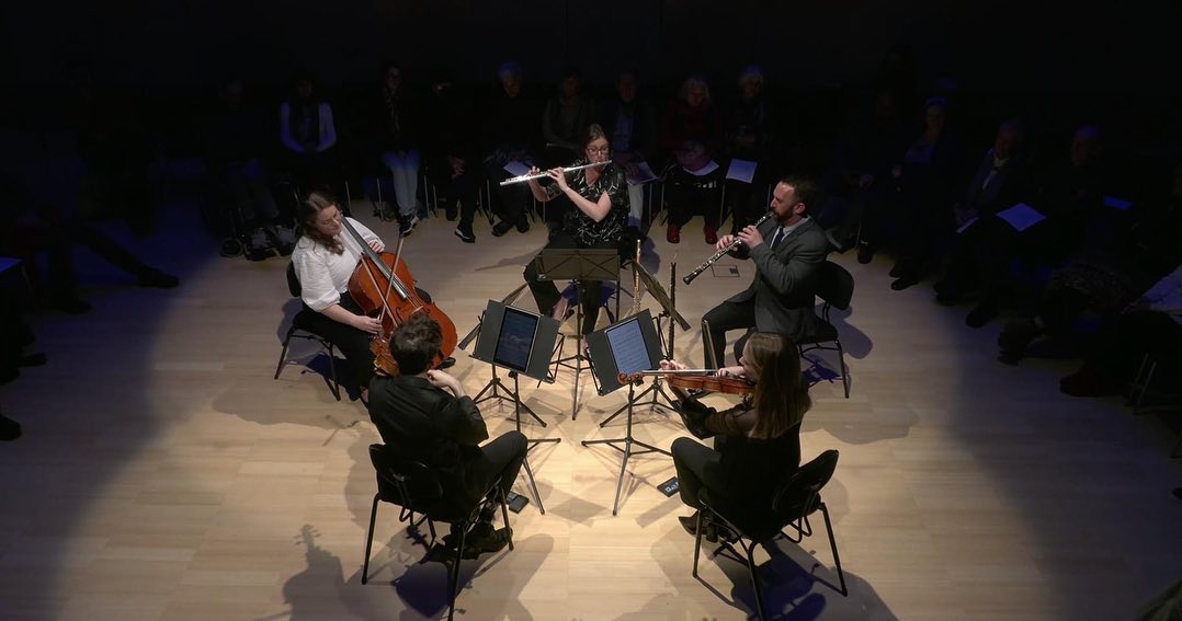 Inventi Ensemble in performance, seated in a circle illuminated by stage lighting with audience members observing on all sides in shadow